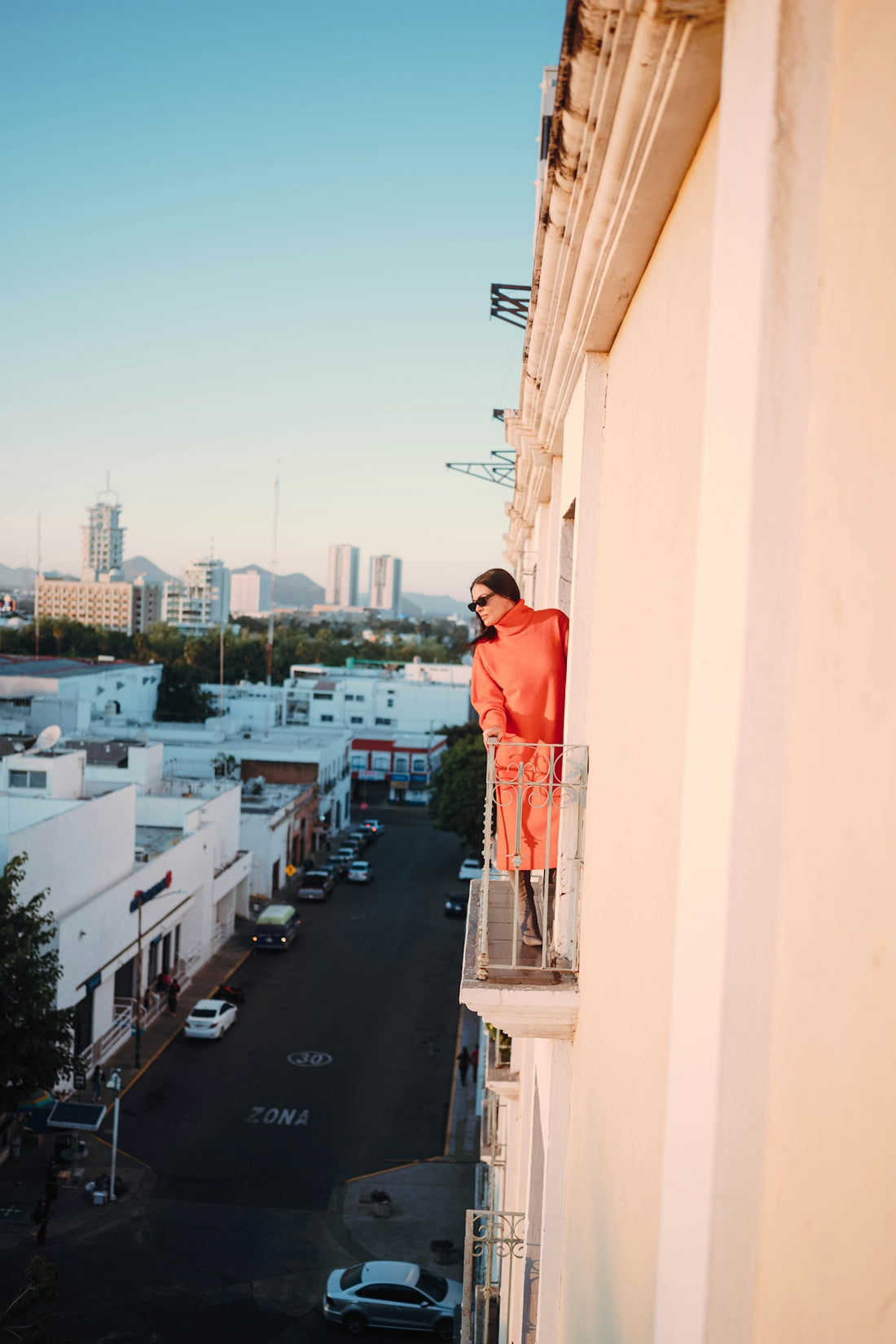 Woman in orange outfit on balcony overlooking city street.